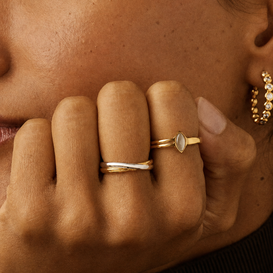 A close-up of a person's hand wearing two gold rings, one with a twisted band and the other with a marquise-cut clear stone, against a warm brown background.